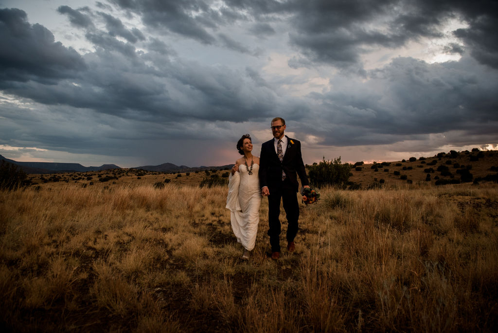 Couple walking through the West Texas expanse surrounded by storm clouds overhead as they look at each other, the groom holding a bouquet of peach roses and greenery photo by Jamie Hardin Houston Wedding Photographer
