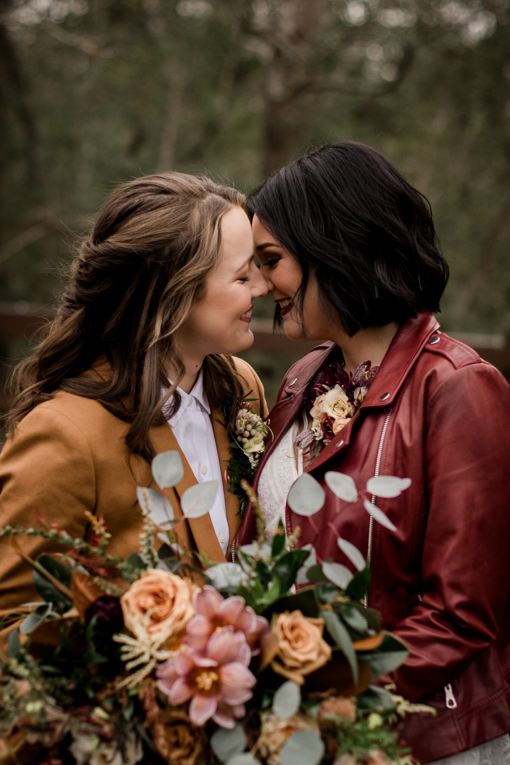 Same sex couple smiling at each other holding a pink and peach colored bouquet, one with light brown hair wearing a mustard jacket and white shirt and the other with raven colored hair wearing a cranberry colored jacket over a white wedding gown photo by Wedding Photographer in Houston, Jamie Hardin Photography