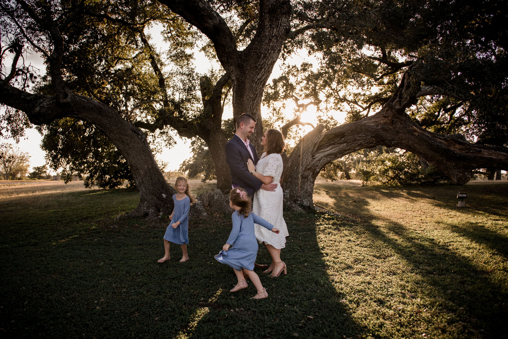 two girls in light blue sundresses running around their mom in a white dress and dad in a blue suit under a massive oak tree and the sun peering from behind the leaves of the massive oak Houston Family Photographer Jamie Hardin Photography