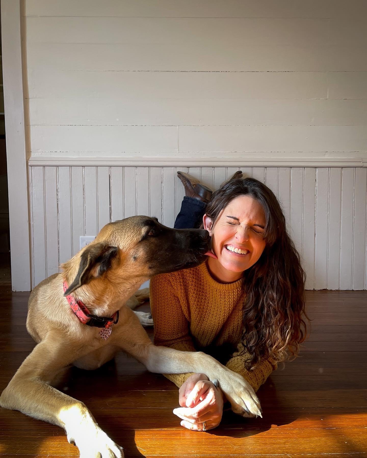 Houston Wedding Photographer Jamie Hardin laying on the floor with her light tan and black dog licking her cheek as she smiles