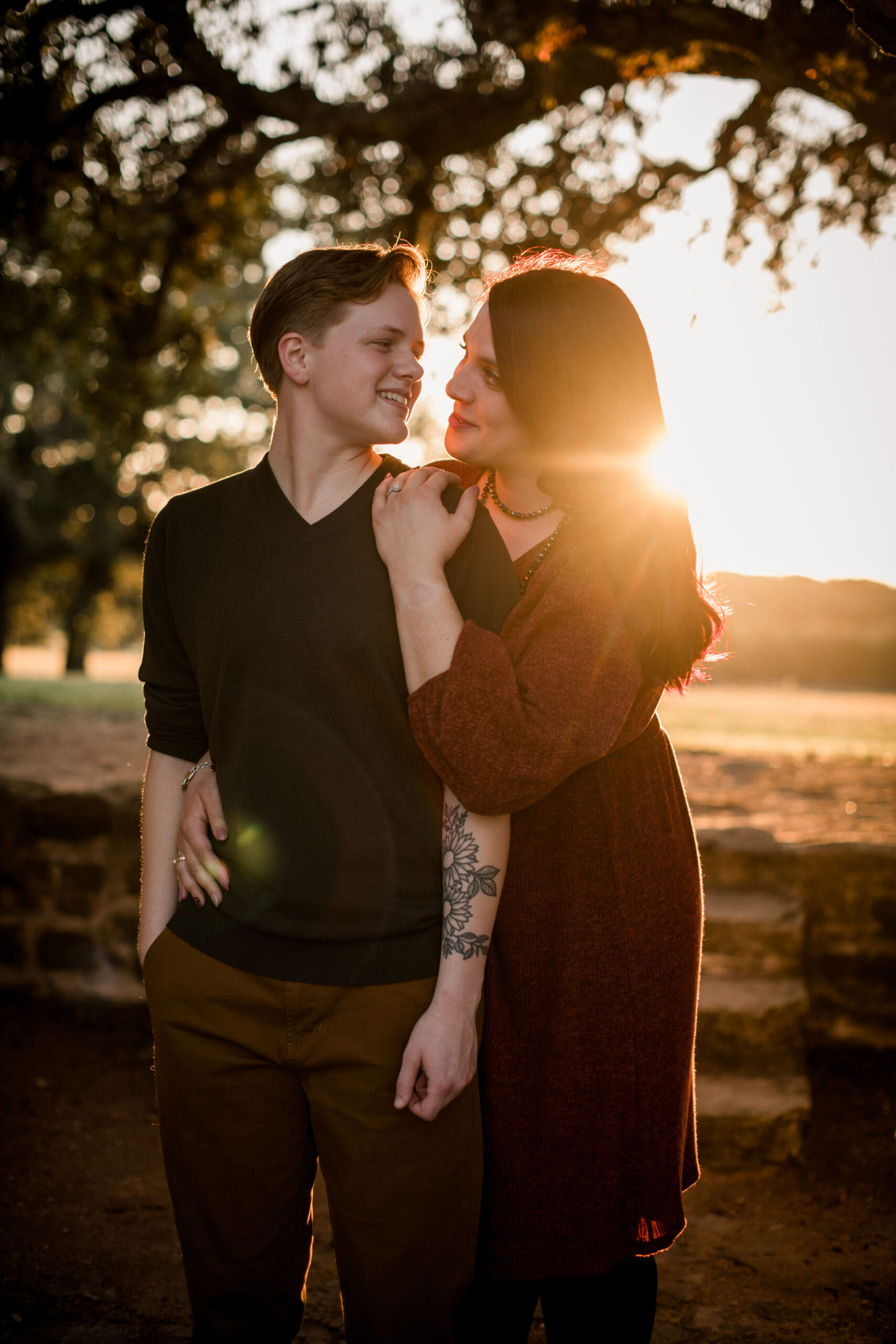 LGBTQ+ couple posing, one wearing a black V tee shirt and khakis with short hair while the other person stands and leans on them draping a hand over their shoulder and wearing a maroon long sleeved linen dress and the sun setting behind them, photo by Same Sex Houston Photographer Jamie Hardin
