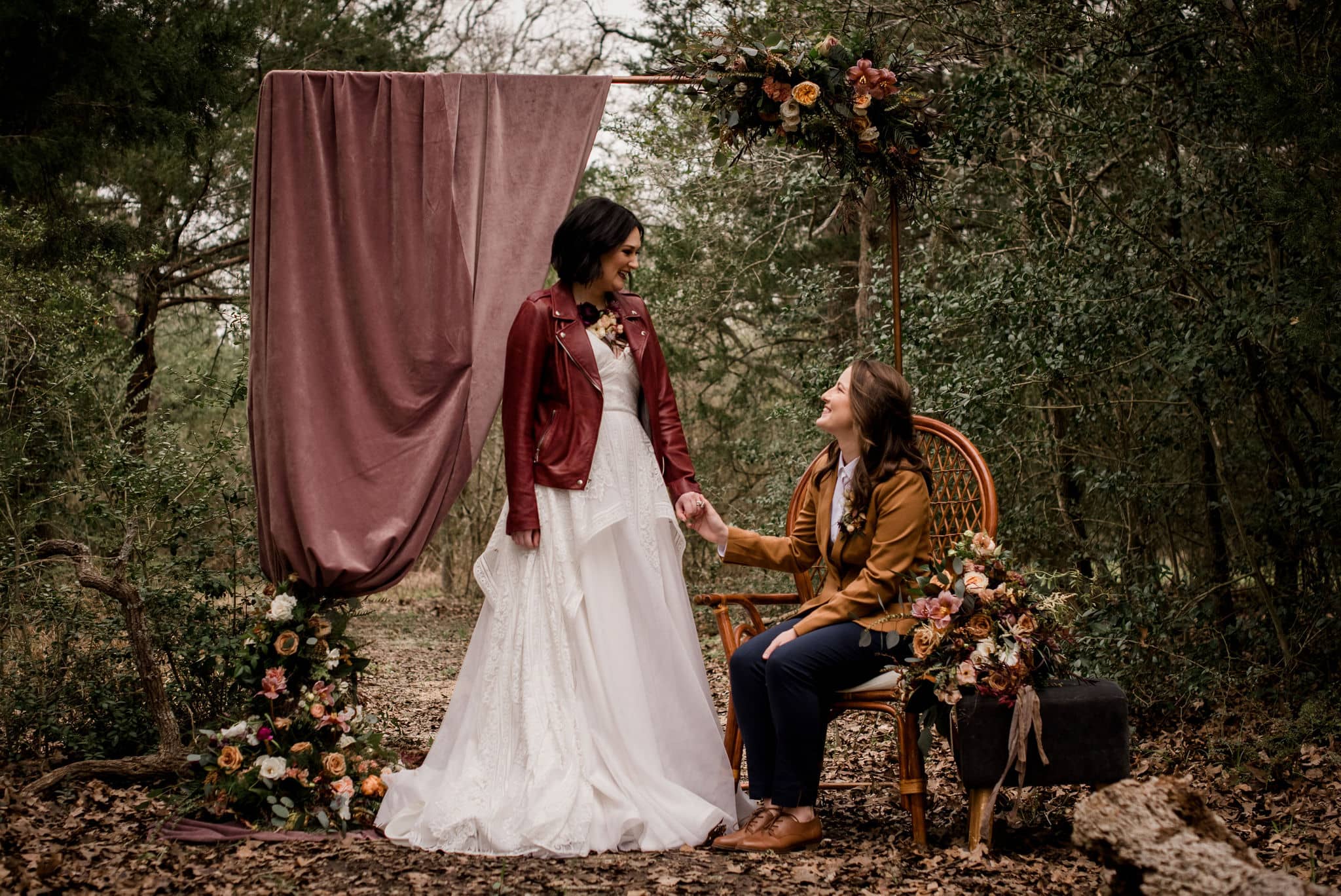 at the back courtyard of peach creek ranch, the texas brides are posed with a decorative backdrop of a mauve arch. the texas bride in a mustard yellow suite is seated while the texas bride in a red jacket and white dress is holding her hand