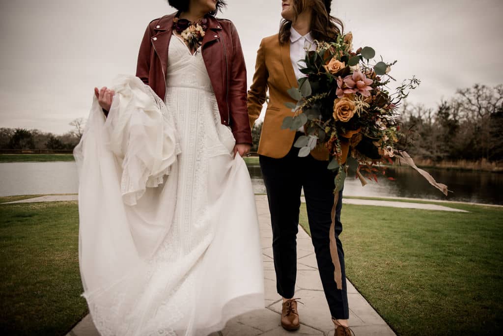 the texas brides are holding hands at the back of peach creek ranch. the courtyard overlooks the lake and the brides are in their wedding attire of a white dress and mustard yellow tuxedo