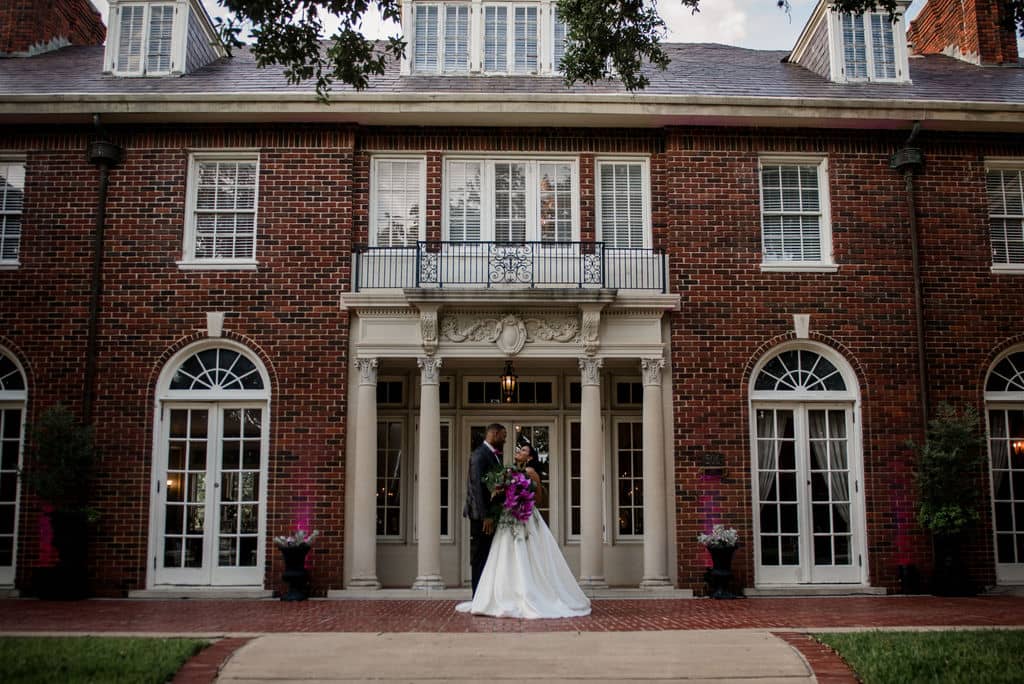 the texas bride and groom share a kiss at the front door steps of the Astin Mansion in Bryan Texas. the mansion is a giant red brick building witih white pillars holding up the front door