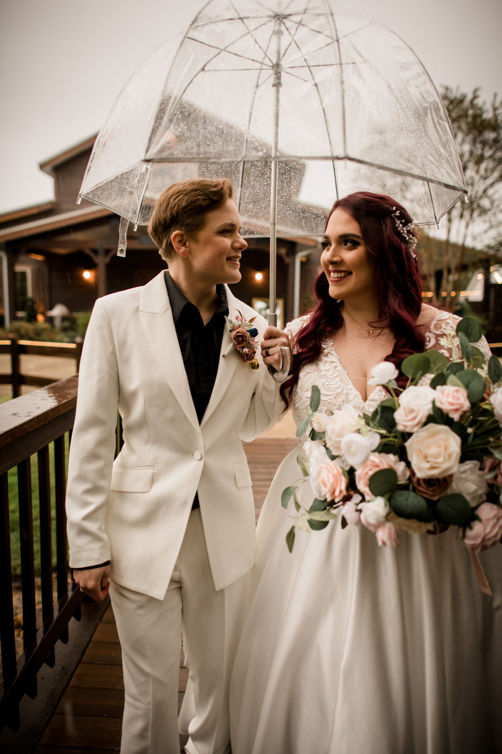 Same sex couple smiling at each other holding a pink and peach colored bouquet, one with light brown hair wearing a mustard jacket and white shirt and the other with raven colored hair wearing a cranberry colored jacket over a white wedding gown photo by Wedding Photographer in Houston, Jamie Hardin Photography