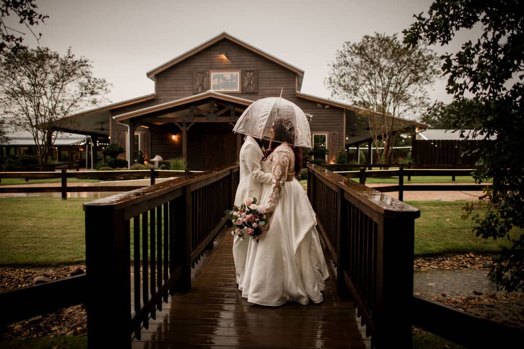 the texas brides pose at the front of the walk way of Peach Creek Ranch kissing under an umbrella in the texas rain. this is a ranch style wedding in the Bryan-College Station area