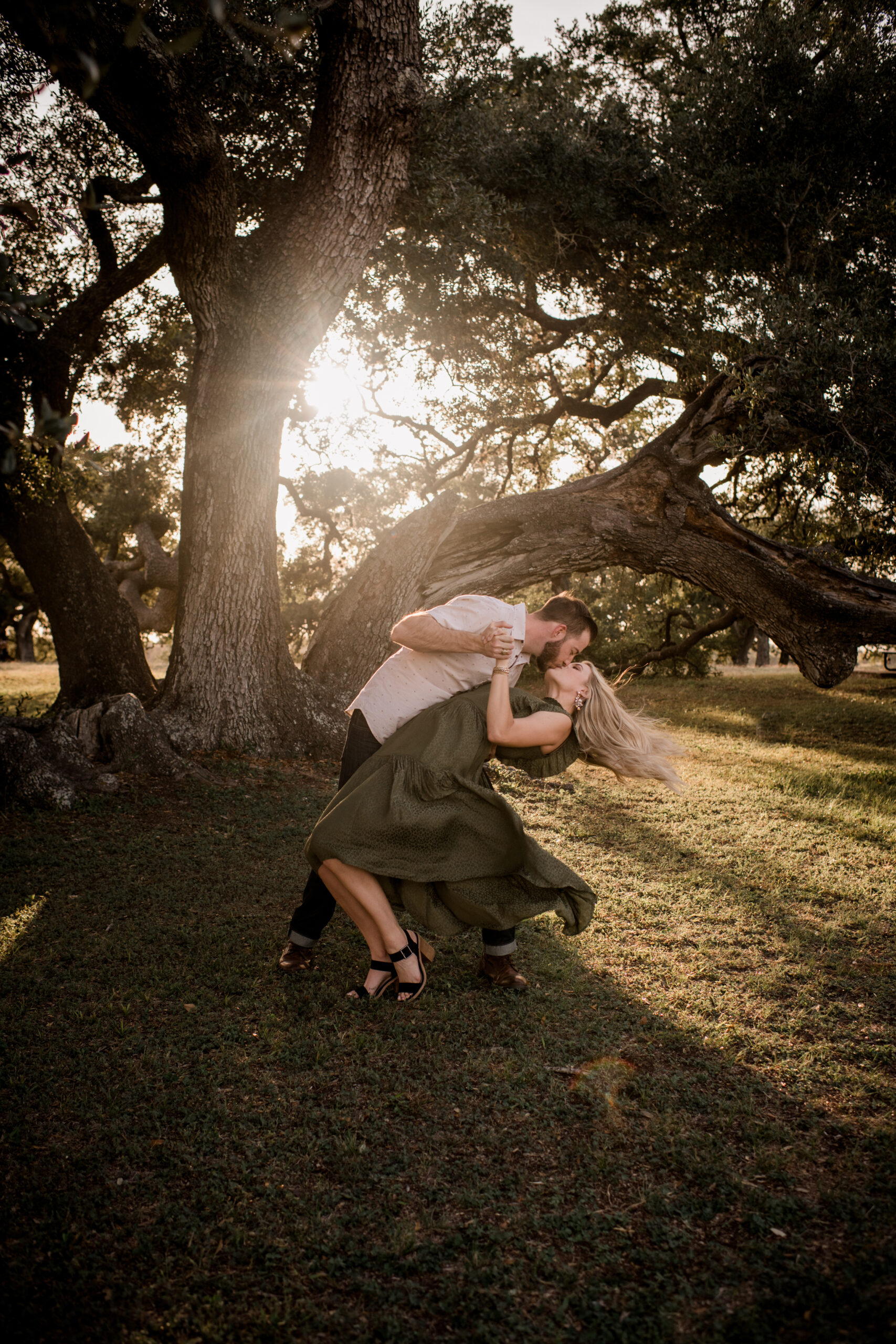 LGBTQ+ couple posing, one wearing a black V tee shirt and khakis with short hair while the other person stands and leans on them draping a hand over their shoulder and wearing a maroon long sleeved linen dress and the sun setting behind them, photo by Same Sex Houston Photographer Jamie Hardin