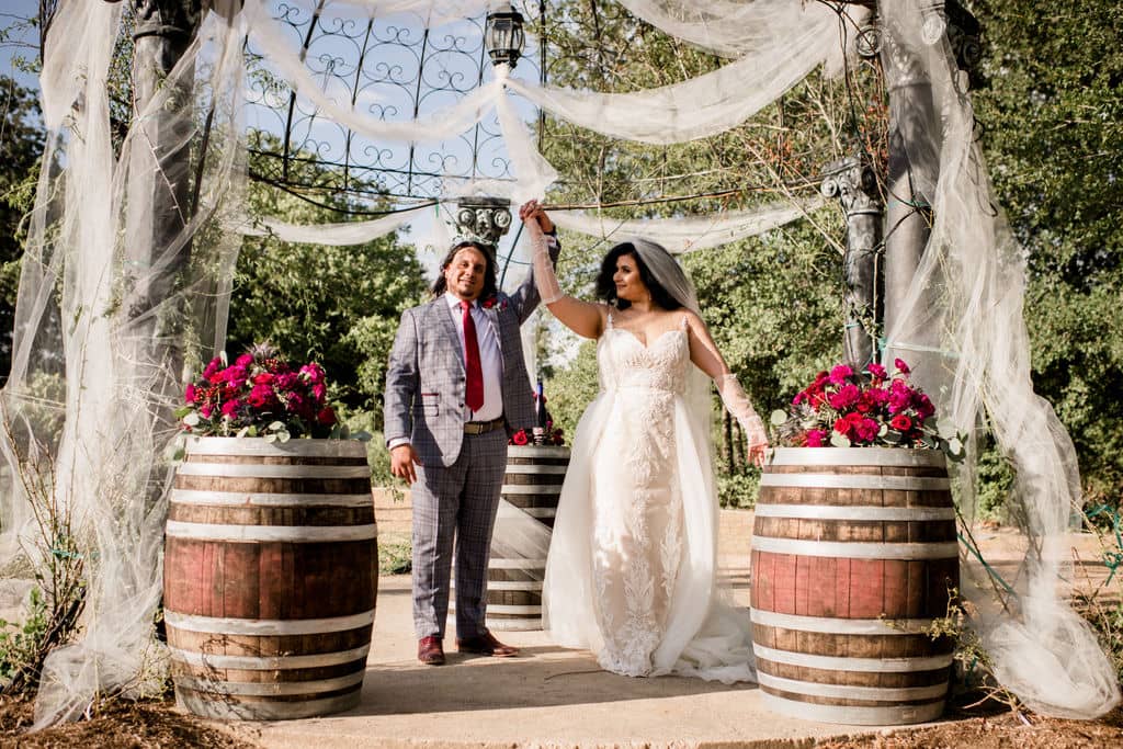 the bride and groom raise their hands in happiness after eloping at texas's messina hof winery's barrel room