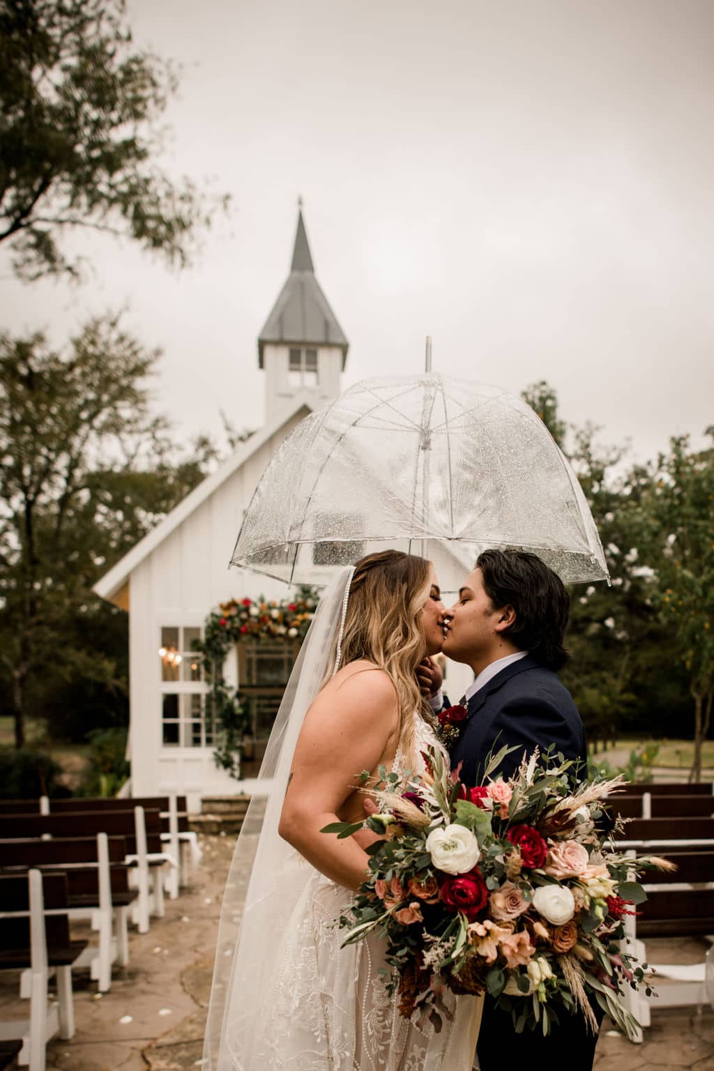 bride and groom kissing in front of the popular white chapel of 7F Lodge and Events in College Station, texas under the rain