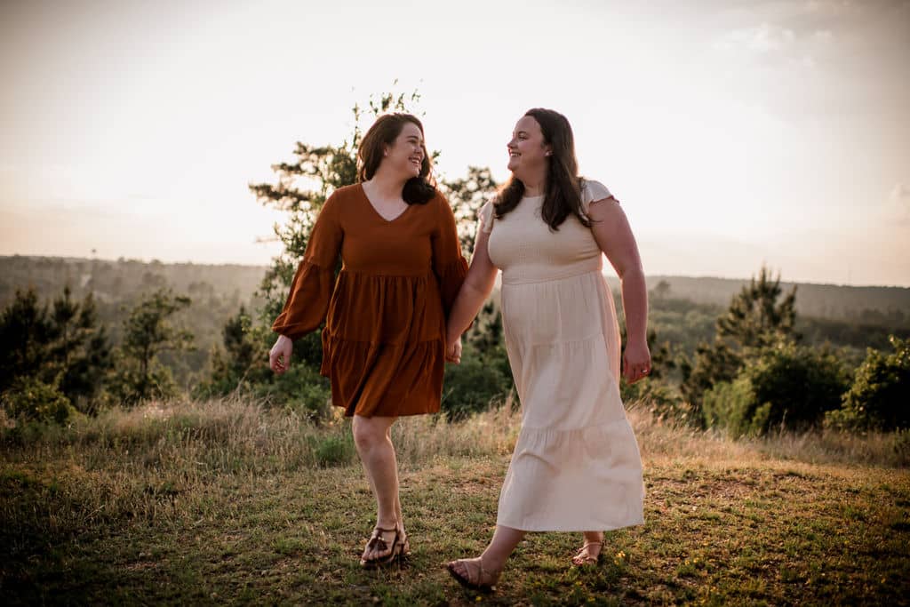 the future brides are holding hands walking in the field in their engagement photo outfits