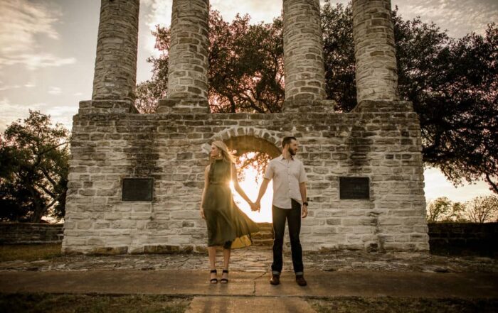 the couple stands in front of a brick structure holding hands facing away from each other in their engagement photo outfits