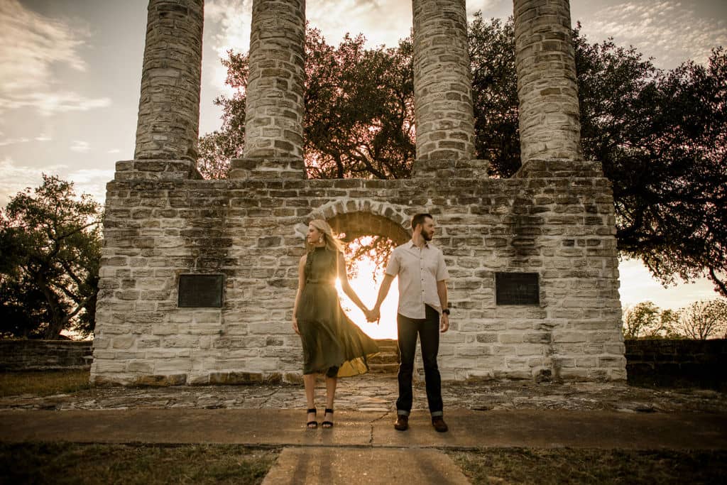 the couple stands in front of a brick structure holding hands facing away from each other in their engagement photo outfits