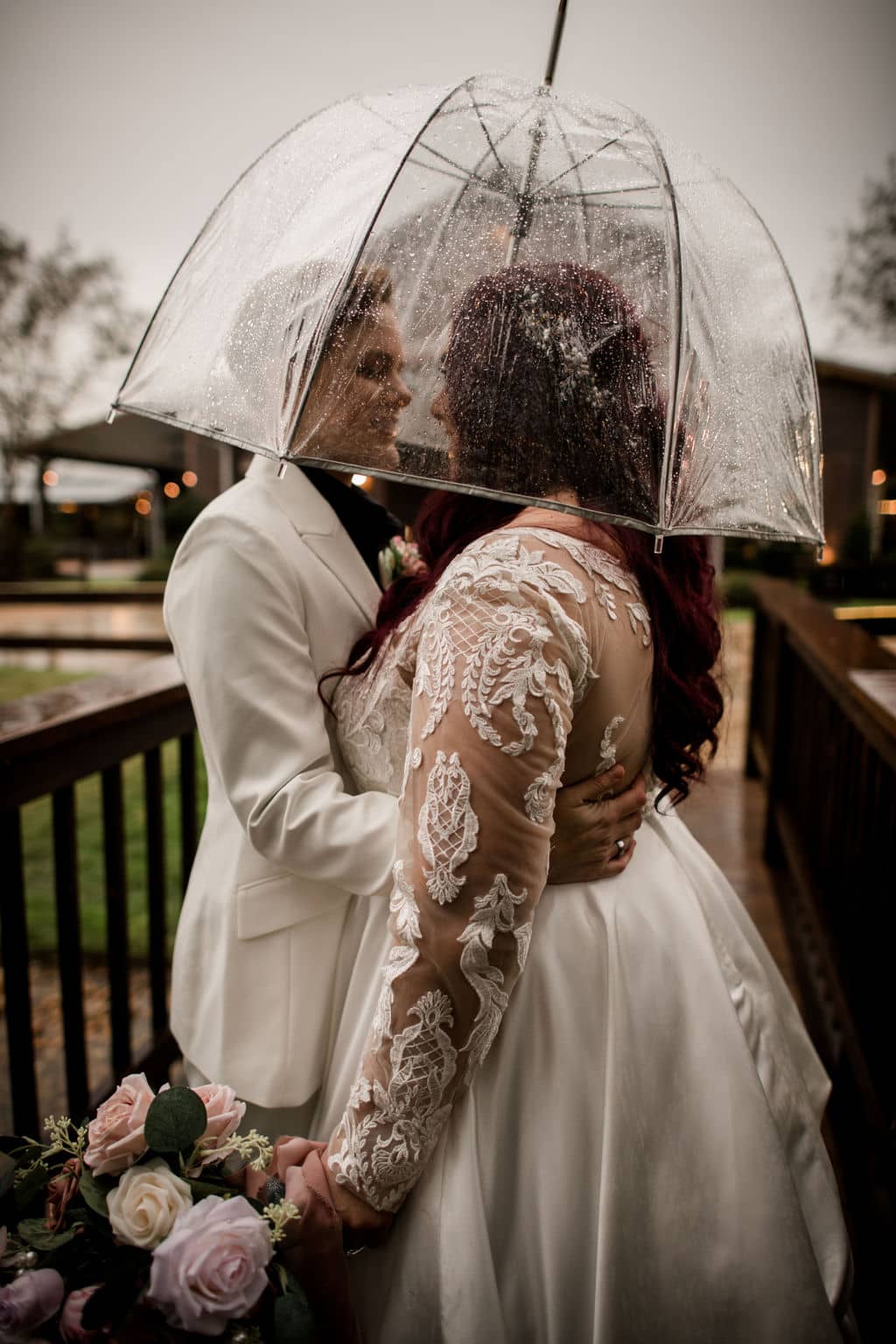 the texas brides embrace each other in white under a clear umbrella