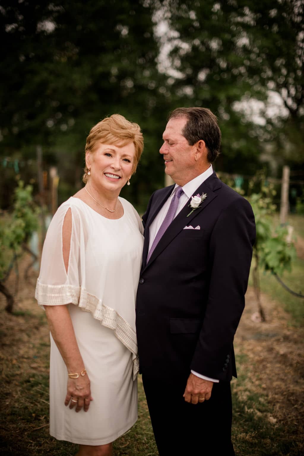 while the wife is looking into the camera, the husband is looking at her on their vow renewal day in a texas vineyard